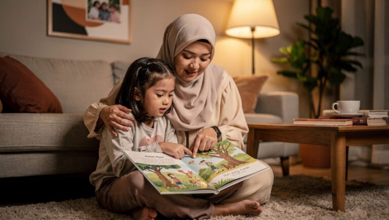 Ibu Muslim berjilbab sedang membacakan buku dan memeluk anaknya, ilustrasi cara menjelaskan musibah dan kematian kepada anak dengan lembut.