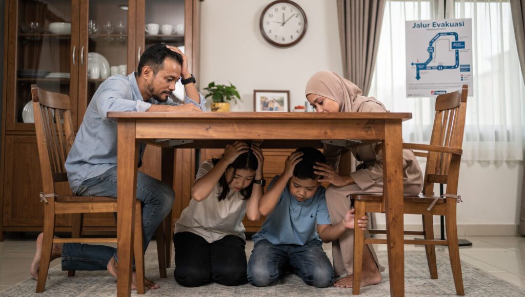 Keluarga muslim melakukan simulasi tindakan penyelamatan diri "drop, cover, and hold on" di bawah meja saat latihan gempa bumi di rumah.